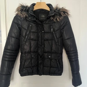 Svart pufferjacka med fuskpäls  - Säljer en svart pufferjacka med avtagbar huva och fuskpäls. Jackan har dragkedja, knappar, två bröstfickor med dragkedja och två fickor med lock framtill. Quiltad design och långärmad modell.
