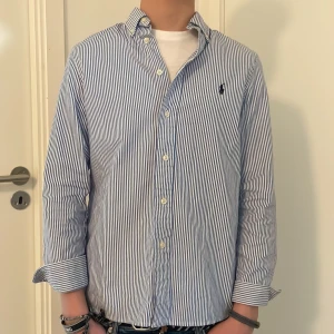 Blårandig skjorta från Polo Ralph Lauren - Snygg blårandig skjorta från Polo Ralph Lauren i slim fit-modell. Skjortan har klassisk krage, vita knappar och broderad logga på bröstet. Tillverkad i bomull och passar perfekt till en stilren look och är använd ett par fåtal gånger.