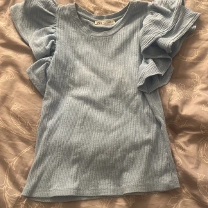 Ljusblå ribbad topp med volangärmar från Zara - Supersöt ljusblå topp från Zara med ribbad struktur och stora volangärmar som ger en trendig look. Perfekt för dig som vill sticka ut lite extra med detaljerade ärmar. Mjuk och bekväm att bära. Använder inte längre då jag inte gillar den, tycker inte den passar mig tar bara Swish 