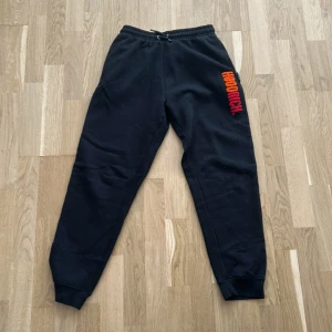 Svarta mjukisbyxor från Hoodrich - Svarta mjukisbyxor från Hoodrich med resår i midjan och mudd vid benslut. Byxorna har en färgglad Hoodrich-logga i orange och rött på vänster lår. Perfekta för en avslappnad stil.
