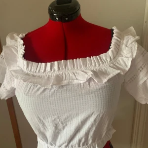 Vit blus med volang från H&M - Säljer en söt vit blus från H&M med bred volang runt halsringningen och korta puffärmar. Blusen har en smockad resår i midjan och är tillverkad i ett lätt bomullstyg. Perfekt för vår och sommar!
