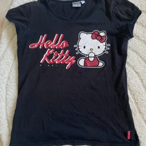 Svart Hello Kitty t-shirt - Svart t-shirt med stort Hello Kitty-tryck framtill och text i rött och vitt. Klassisk rund halsringning och korta ärmar. 💕