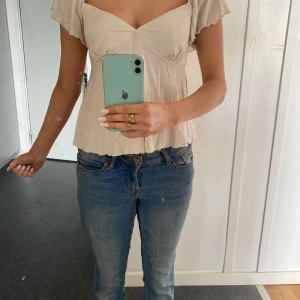 Beige omlottopp med knytband - Supersöt beige omlottopp med v-ringning och korta volangärmar. Toppen har knytband i ryggen och är tillverkad i ett mjukt, tunt material som ger en luftig känsla. Perfekt för en avslappnad och trendig stil.