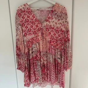 Mönstrad rosa och röd klänning från Zara - Supersöt klänning från Zara i rosa och röda toner med blommigt och grafiskt mönster. Långärmad modell med v-ringning och volangdetaljer framtill. Perfekt för vår och sommar!