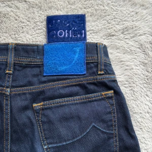Mörkblå jeans från Jacob Cohen - Säljer ett par mörkblå jeans från Jacob Cohen med klassisk femficksdesign och kontrastsömmar. Jeansen har en snygg blå patch bak i midjan och raka ben. Perfekta för en stilren och avslappnad look. Storlek 30 skulle säga att dom är lite mindre i storleken och passar dig som är 170-175