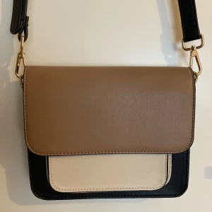 Beige och svart axelväska  - Säljer en stilren axelväska i beige, svart och krämvitt. Väskan har ett lock i beige, svart kropp och en krämvit ficka framtill. Justerbar axelrem med guldfärgade metalldetaljer. 