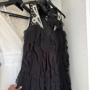 Svart festklänning med pärlor och fjädrar - Supersnygg svart klänning med volanger, pärldetaljer och fjädrar vid axlarna. Ärmlös modell med rund halsringning och dekorativa detaljer som ger en lyxig känsla. Perfekt för dig som vill sticka ut på festen!