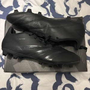 Svarta Adidas Predator fotbollsskor - Säljer ett par helsvarta fotbollsskor från Adidas med snörning och rund tå. Storlek 45 och 1/3 Skorna har en syntetisk ovandel och mönstrad sula med dobbar för bra grepp på planen. Perfekta för dig på fotbollsplanen. Dojorna är aldrig använda! Helt nyskick, inga skavanker ingenting. Priset är förhandlingsbart! Lådan medföljer Bara att skriva om ni undrar över något😁🤩👍 