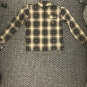 Svart och grå rutig skjorta med stjärnmönster från Dope - Säljer en långärmad skjorta från Dope i svart och grått med rutigt mönster och små stjärnor. Skjortan har en bröstficka och klassisk krage. Perfekt för dig som gillar streetwear och vill sticka ut lite extra. SOFFFT