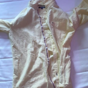 Gul skjorta från Tommy Hilfiger - Ljusgul skjorta från Tommy Hilfiger med klassisk krage och knappar framtill. Skjortan har lång ärm och en bröstficka med liten logga. Tillverkad i mjuk bomull och har diskreta Hilfiger-detaljer vid knappslån och ryggen.