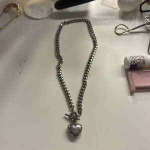 Chunky silver halsband med hjärta - Snyggt chunky halsband i silverfärgad metall med ett stort hjärtformat hänge. Kedjan är grov och har ett coolt T-lås som detalj. Perfekt för dig som vill ha ett statement piece med lite edge och romantisk vibe. Bara använt ett fåtal gånger.