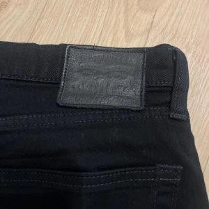 Svarta Levi's jeans straight fit - Svarta jeans från Levi's med klassisk röd logotagg vid bakfickan och svart patch i midjan. Modellen har raka ben och fem fickor, med snygga svarta sömmar och metallknappar. Perfekt för dig som gillar stilrena och tidlösa jeans.