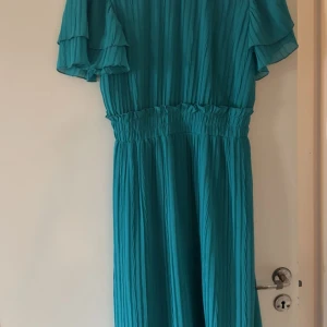 Turkos plisserad långklänning från Zara - Säljer en turkos långklänning från Zara med plisserat tyg och volangdetaljer på de korta ärmarna. Klänningen har markerad midja med resår och en snygg v-ringad hals. Perfekt för dig som gillar färg och flowy vibes. 549