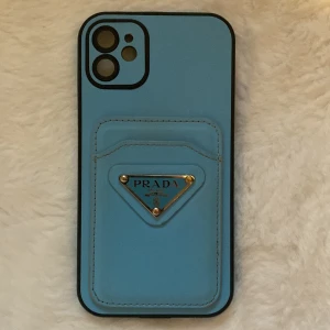 Blå Prada mobilskal - Snygg ljusblå mobilskal från Prada med inbyggd korthållare och ikonisk metallogo framtill. Väskan är gjord i skinn och har svarta kanter som ger en clean kontrast. Perfekt för dig som vill ha både stil och funktion i ett. Passar iPhone 11