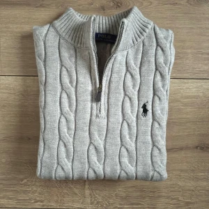 Grå stickad tröja Polo Ralph Lauren - Säljer en grå stickad tröja från Polo Ralph Lauren med klassiskt flätmönster och half zip vid halsen. Tröjan har ribbad krage och det ikoniska Polo-logot broderat på bröstet. Perfekt för dig som gillar stilren och tidlös design.