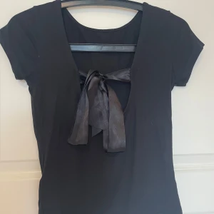 Svart topp med rosett - Svart kortärmad topp, en snygg rosett baktill. Materialet är mjukt och stretchigt. Rosetten är i ett glansigt tyg som ger en cool kontrast mot resten av toppen. Perfekt för dig som gillar stilrena detaljer.