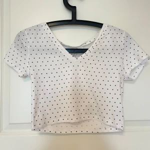 Vit croppad prickig topp med v-ringning - Supersöt croppad topp i vit med svarta prickar. V-ringad framtill och rundad rygg, korta ärmar och mjukt bomullsmaterial. Perfekt att styla med jeans eller kjol för en chill och trendig look.