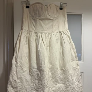 Vit axelbandslös broderad klänning - Säljer en vit axelbandslös klänning med markerad byst och vackra broderade detaljer nertill. Klänningen har en utsvängd kjol och är perfekt för dig som gillar romantisk och feminin stil. Materialet känns som bomull och broderierna ger en lyxig känsla.