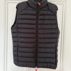 Svart dunväst från Save The Duck - Säljer en svart dunväst från Save The Duck i storlek L. Använt 1 gång. Västen är quiltad med horisontella sömmar, har hög krage och hel dragkedja med orange detaljer. 