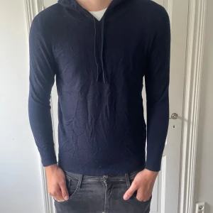 Mörkblå kashmirhoodie från Sayless - Mörkblå hoodie i mjuk kashmir från Sayless. Tröjan har lång ärm, huva med snörning och ribbade muddar. Snygg och enkel modell som är perfekt när du vill vara både chill och stilren.