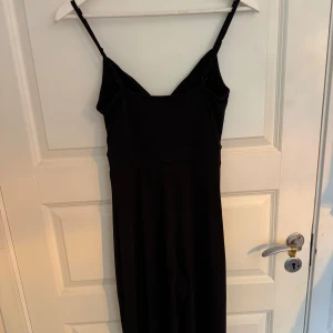 Svart jumpsuit från H&M Divided - En stilren svart jumpsuit från H&M Divided med smala axelband och rak passform. Jumpsuiten är ärmlös och har en enkel, elegant design som passar perfekt till många olika tillfällen. Materialet känns mjukt och följsamt.