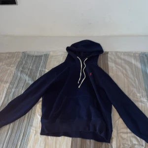 Marinblå hoodie från Polo Ralph Lauren - Snygg marinblå hoodie från Polo Ralph Lauren med vit snörning och klassisk känguruficka. Liten röd logga broderad på bröstet. Perfekt för dig som gillar stilrena och bekväma plagg med premiumkänsla.