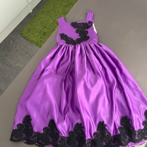 Lång lila klänning med svarta spetsdetaljer - Säljer en elegant lång lila klänning med breda axelband och svarta broderade spetsdetaljer både upptill och längs nederkanten. Kjolen är voluminös och har en lyxig satinliknande finish som ger en riktigt festlig känsla.