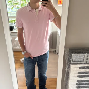 Ljusrosa pikétröja med krage - Säljer en ljusrosa pikétröja med vit krage och knappslå. Tröjan har korta ärmar med vit kant och en diskret broderad logga på bröstet. Perfekt för dig som gillar stilrena och softa färger. Materialet känns som bomull och är skönt mot huden.