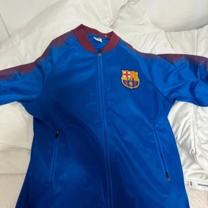 Blå FC Barcelona vindjacka - Snygg blå vindjacka från FC Barcelona med klubbens emblem på bröstet. Jackan har dragkedja framtill, två fickor med dragkedja och vinröda detaljer vid krage och axlar. Perfekt för dig som älskar fotboll och vill visa vilket lag du hejar på. Köpte tröjan på en marknad i Stockholm för 300 och har använt den en gång. Pris kan diskuteras 