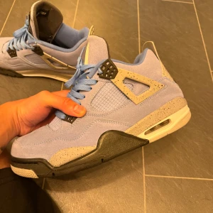 Nike Air Jordan 4 University Blue - Nike Air Jordan 4 University Blue med ljusblå mocka, svarta och grå detaljer samt stänkmönstrad mellansula. Klassisk meshpanel på sidorna, blå skosnören och ikonisk Jumpman-logga på innersulan. Perfekt för dig som vill sticka ut med stilrena sneakers.