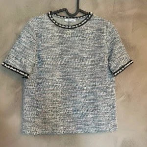 Tweedblus med pärldetaljer Zara Woman - Säljer en snygg tweedblus från Zara Woman i blått och vitt med kort ärm. Blusen har svarta och silvriga kanter med pärlor vid halsringning och ärmslut. Rak passform och klassisk rund hals. Perfekt för dig som gillar unika detaljer!