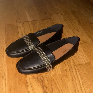 Svarta loafers med nitar från Topshop - Snygga svarta loafers från Topshop med fyrkantig tå och bred rem dekorerad med silvriga nitar. Skorna är i skinnimitation och har platt sula, perfekt för en trendig och stilren look. Passar till både jeans och kostymbyxor.