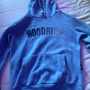 Blå hoodie från Hoodrich - Säljer en blå hoodie från Hoodrich med broderad logga framtill. Tröjan har huva, magficka och långa ärmar. Perfekt för dig som gillar streetwear och vill ha en chill look. Materialet känns mjukt och skönt, troligen bomull eller bomullsblandning.