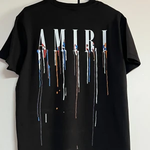 Svart AMIRI t-shirt med färgstänk - Svart t-shirt från AMIRI med stor logga och coolt färgstänksmönster i vitt, blått, orange och rött på både bröstet och ryggen. Klassisk rund hals och korta ärmar. Materialet är mjuk bomull som känns skönt mot huden. Perfekt för dig som gillar streetwear och statement-plagg.