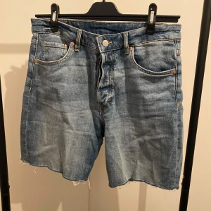 Blåa bermuda jeansshorts &Denim - Snygga blå bermuda jeansshorts från &Denim med hög midja och råa kanter. Klassisk femficksmodell med knappgylf och slitna detaljer för en avslappnad vibe. Perfekta till sommaren och lätta att matcha med allt.
