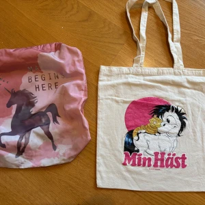 Två tygväskor med hästmotiv - Säljer två tygväskor: en rosa shoppingväska med enhörning och texten 'Magic begins here', samt en beige shoppingväska med tryck från Min Häst och rosa cirkel. Perfekta för skolan eller stan, båda med långa handtag och lekfull design. Kan finnas ngn fläck på den rosa 