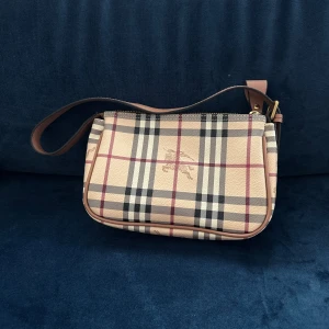 Rutig axelväska från Burberry - Säljer en klassisk axelväska från Burberry med ikoniskt rutigt mönster i beige, svart och rött. Väskan har detaljer i brunt skinn, guldfärgade metalldelar och dragkedja upptill. Perfekt storlek för det viktigaste och snygg att bära över axeln.