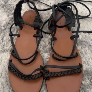 Svarta flätade sandaler med snörning💕 - Snygga sandaler med svarta flätade remmar och långa snören att knyta runt vristen. Sulan är brun och platt, och remmarna är i svart syntetmaterial. Perfekta för sommaren och ger cool vibe till outfitten. Pris kan diskuteras💕