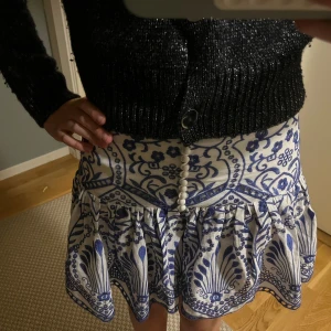 Vit och blå mönstrad kortkjol - Supersöt kortkjol i vitt och blått med unikt mönster och dekorativa knappar framtill. Kjolen har volangkant och en lätt utsvängd form som ger en skön och trendig vibe. Perfekt för dig som vill sticka ut med en cool och fräsch look.