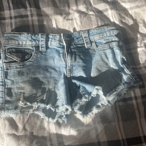 Blå jeansshorts med fransig kant - Snygga ljusblå jeansshorts med fransig och sliten kant. Klassisk femficksmodell med bälteshällor och knappar på bakfickorna. Perfekta för varma dagar och ger en avslappnad vibe.