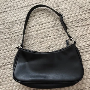 Svart axelväska från Monki i skinnimitation - Stilren svart axelväska från Monki i skinnimitation med justerbart axelband och silverfärgade metalldetaljer. Väskan har dragkedja upptill och en enkel, minimalistisk design som passar till många outfits. Köpt för 2-3 år sedan för 200-300kr. Priset kan diskuteras.