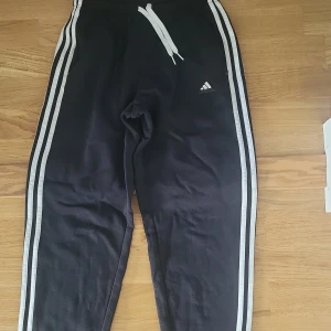 Svarta Adidas mjukisbyxor med vita ränder stl 164 - Svarta mjukisbyxor från Adidas med klassiska vita ränder längs sidorna och Adidas-logga på låret. Byxorna har resår i midjan med vit snörning och muddar vid bensluten. Perfekta för chill eller träning.