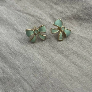 Mintgröna rosettörhängen i guld - Superfina örhängen i form av rosetter med mintgröna detaljer och guldfärgade kanter. Perfekt för dig som vill ha något sött och unikt till din look. Rosettformen ger en lekfull vibe och färgen sticker verkligen ut.