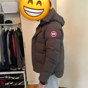 Mörkgrå Mac Millan parka Canada Goose - Säljer en mörkgrå Mac Millan parka  från Canada Goose med huva och klassisk logga på ärmen. Jackan är quiltad, har dragkedja och knäppning framtill samt en clean, stilren look. Perfekt för kalla dagar och riktigt snygg till streetwear.