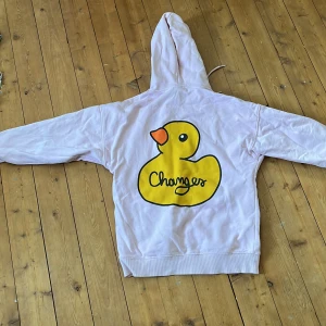 Rosa Bieber hoodie med anka-tryck - Säljer en ljusrosa hoodie från Bieber med stor gul anka och texten 'Changes' på ryggen. Framtill finns en magficka med Bieber-logga i svart. Hoodien har huva med snörning och långa ärmar. Perfekt för dig som gillar coola tryck och streetstyle.