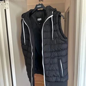 Svart dunväst från Urban Classics - Svart dunväst med huva från Urban Classics. Västen har vita dragkedjor och snören, två fickor framtill och quiltad design. Perfekt för lager-på-lager och chill stil. Materialet är syntet och västen har en loose passform. Använd 1 gång. 