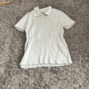 Vit ribbad pikétröja med krage - Säljer en vit ribbad pikétröja från  Lindex med klassisk krage och korta ärmar. Tröjan har tre knappar framtill och är gjord i mjuk bomull. Perfekt för dig som gillar stilrena och bekväma plagg med lite struktur.