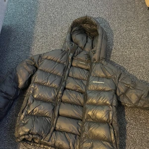 Svart dunjacka Peak Performance M - Säljer en svart dunjacka från Peak Performance i storlek M. Jackan är inte i nyskick och har lite defekter på sig såsom dragkedjorna på fickorna som har öppnat upp sig men borde va lätt att fixa men annars är den i bra skick. Jackan har en rejäl huva, breda quiltade paneler och dragkedja framtill. Perfekt för kalla dagar och riktigt snygg streetstil. Klassisk logga på bröstet och justerbar nederkant.