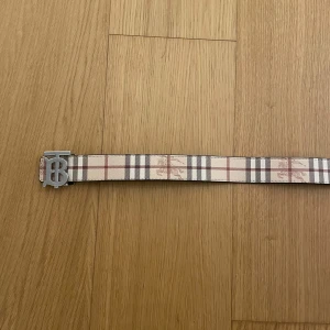 Burberry rutig skärp med silverdetalj - Säljer ett snyggt Burberry-bälte med klassiskt rutigt mönster i beige, brunt och rött. Bältet har en bred design och en stor silvrig spänne med Burberrys ikoniska TB-logo. Perfekt för att lyfta vilken outfit som helst.
