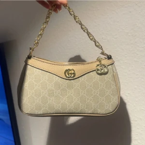 Beige axelväska med kedja - Säljer en beige axelväska med klassiskt GG-mönster och guldfärgad kedja som axelrem. Väskan har detaljer i ljust skinn och dragkedja upptill. Perfekt storlek för det viktigaste och snygg att bära till alla outfits.